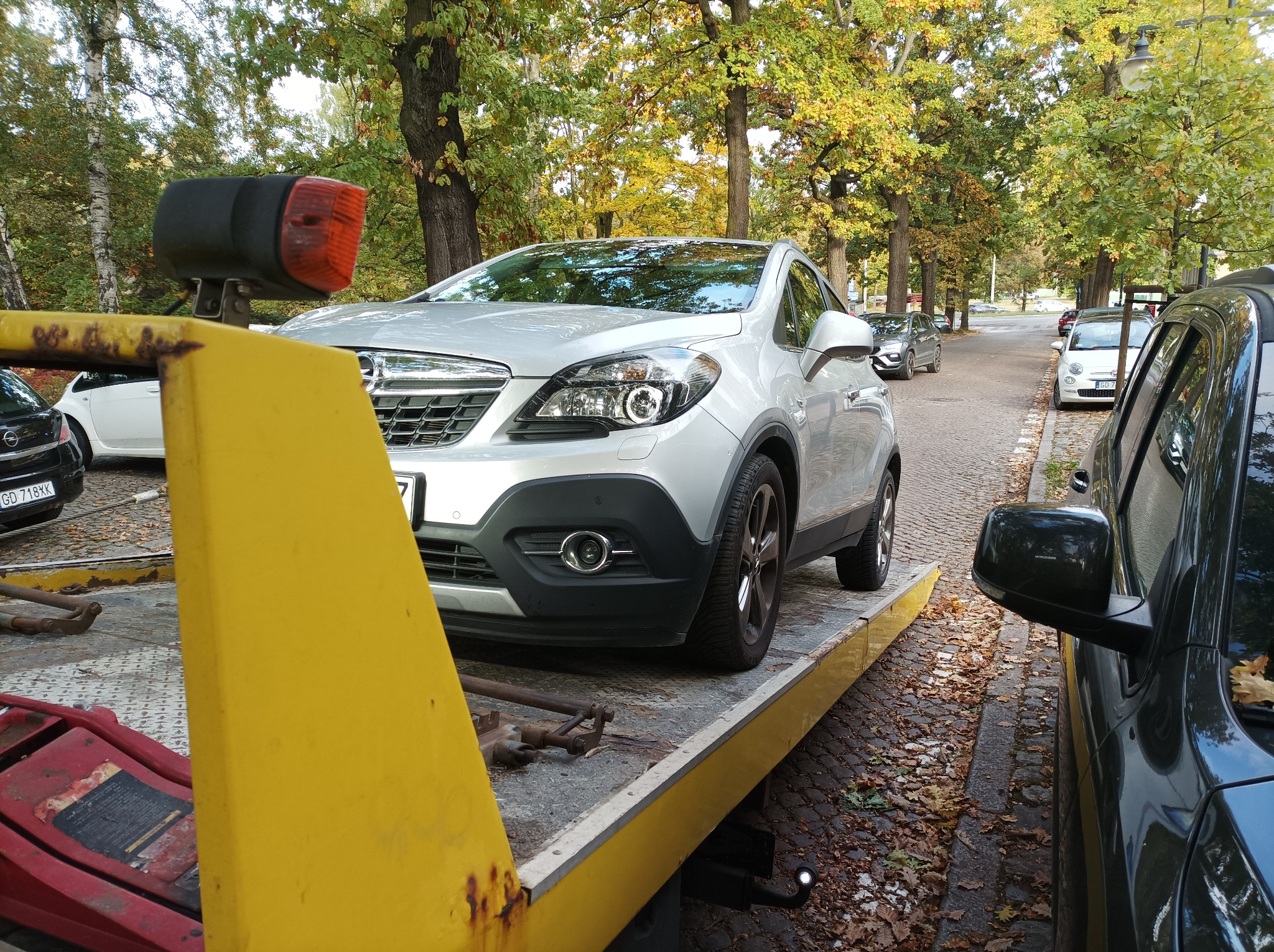 Srebrny Opel Mokka na żółtej lawecie z widocznym reflektorem i narzędziami, w tle jesienny krajobraz parkowy.