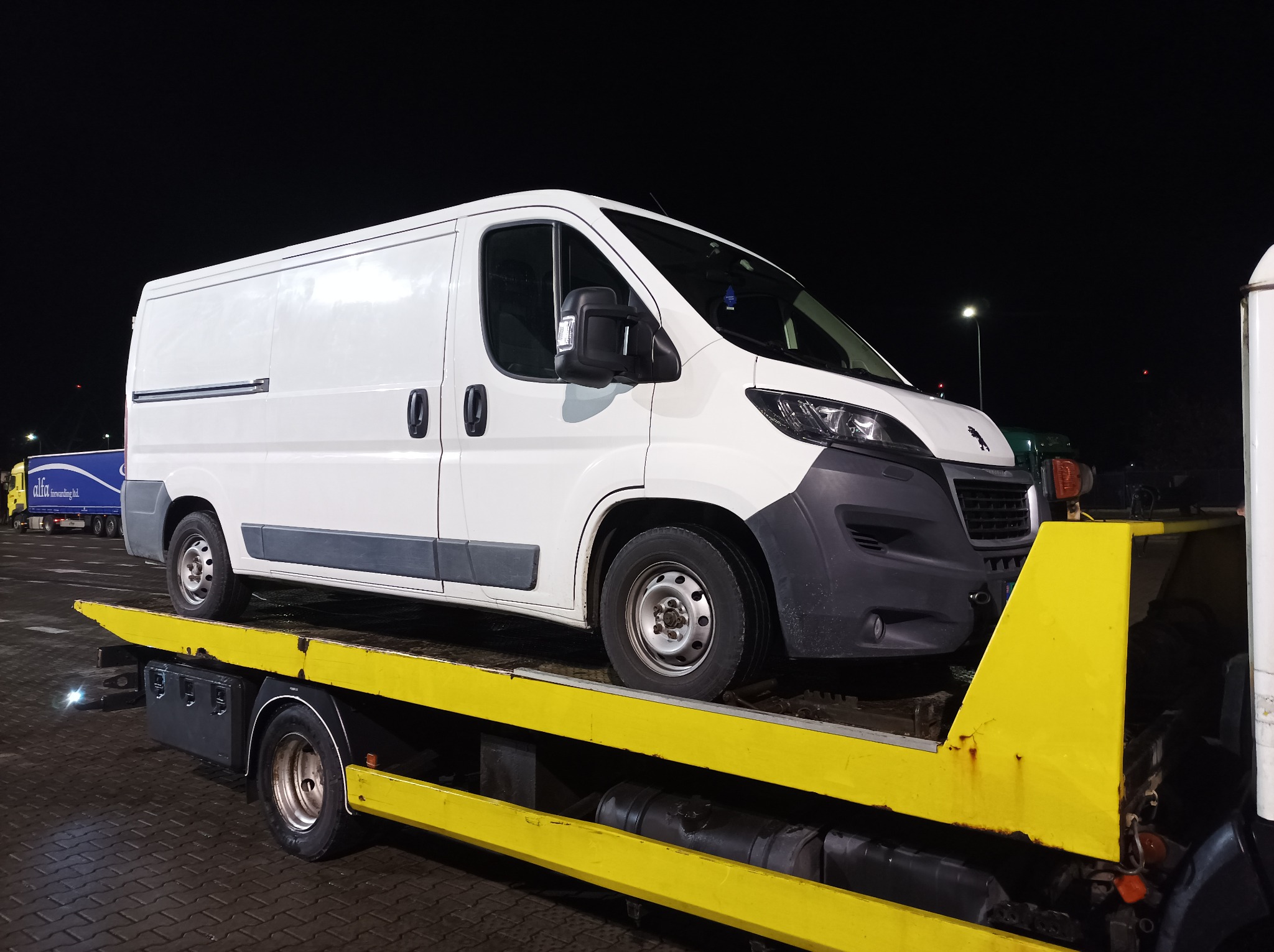 Biały furgon na żółtej platformie transportowej w nocnym otoczeniu, widoczny fragment innego pojazdu w tle.