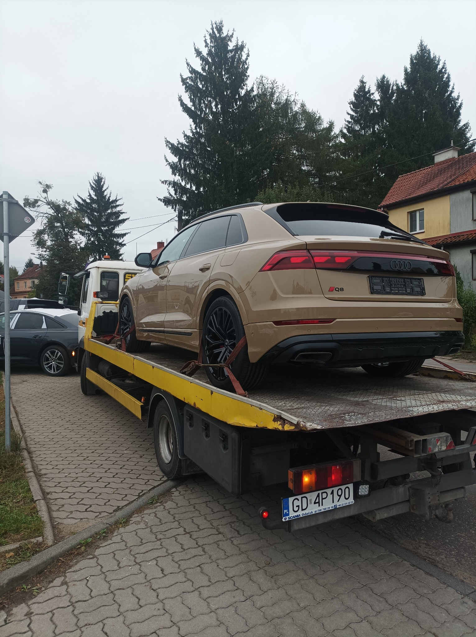 Złoty SUV Audi Q8 zabezpieczony pasami transportowymi na żółtej platformie pojazdu pomocy drogowej z widocznymi światłami ostrzegawczymi, na tle chodnika i zabudowań.