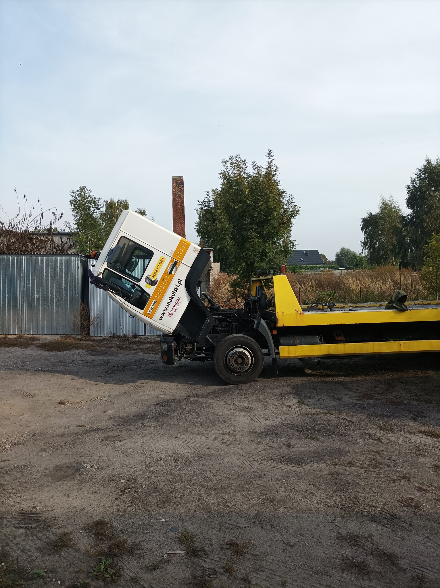 Żółto-biała ciężarówka z podniesioną kabiną, zaparkowana na grząskim terenie, z widoczną ceglaną konstrukcją w tle.