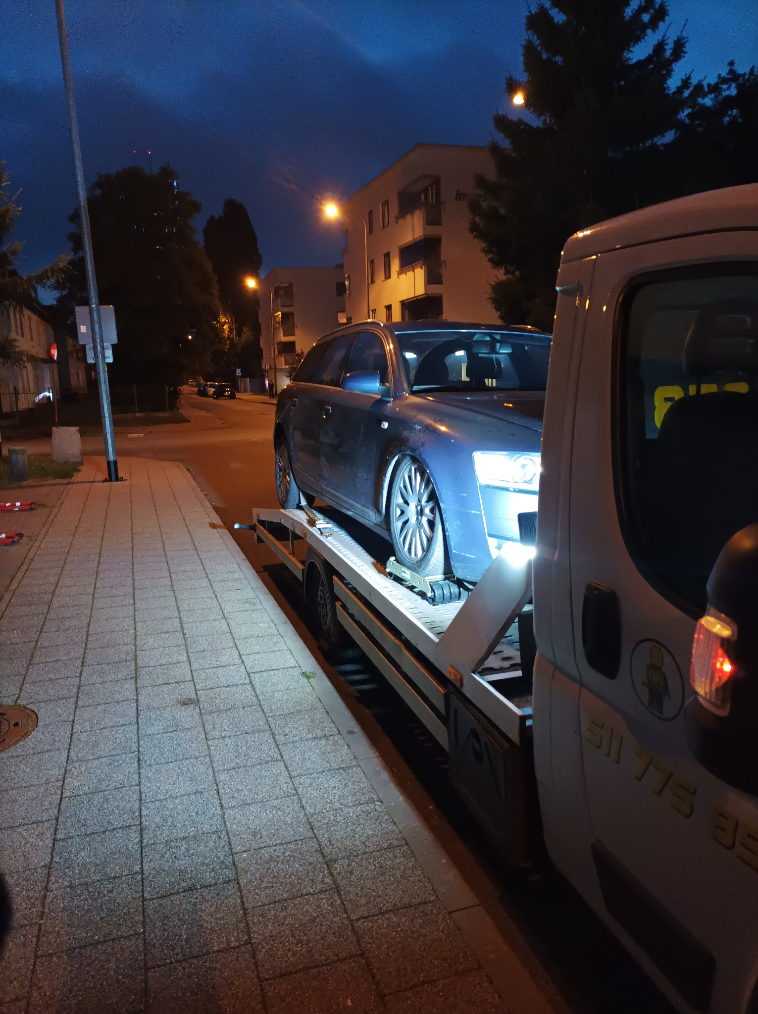 Niebieski samochód osobowy, prawdopodobnie Audi, z uszkodzonym zawieszeniem, załadowany na platformę pojazdu pomocy drogowej na tle chodnika i budynków mieszkalnych po zmroku.