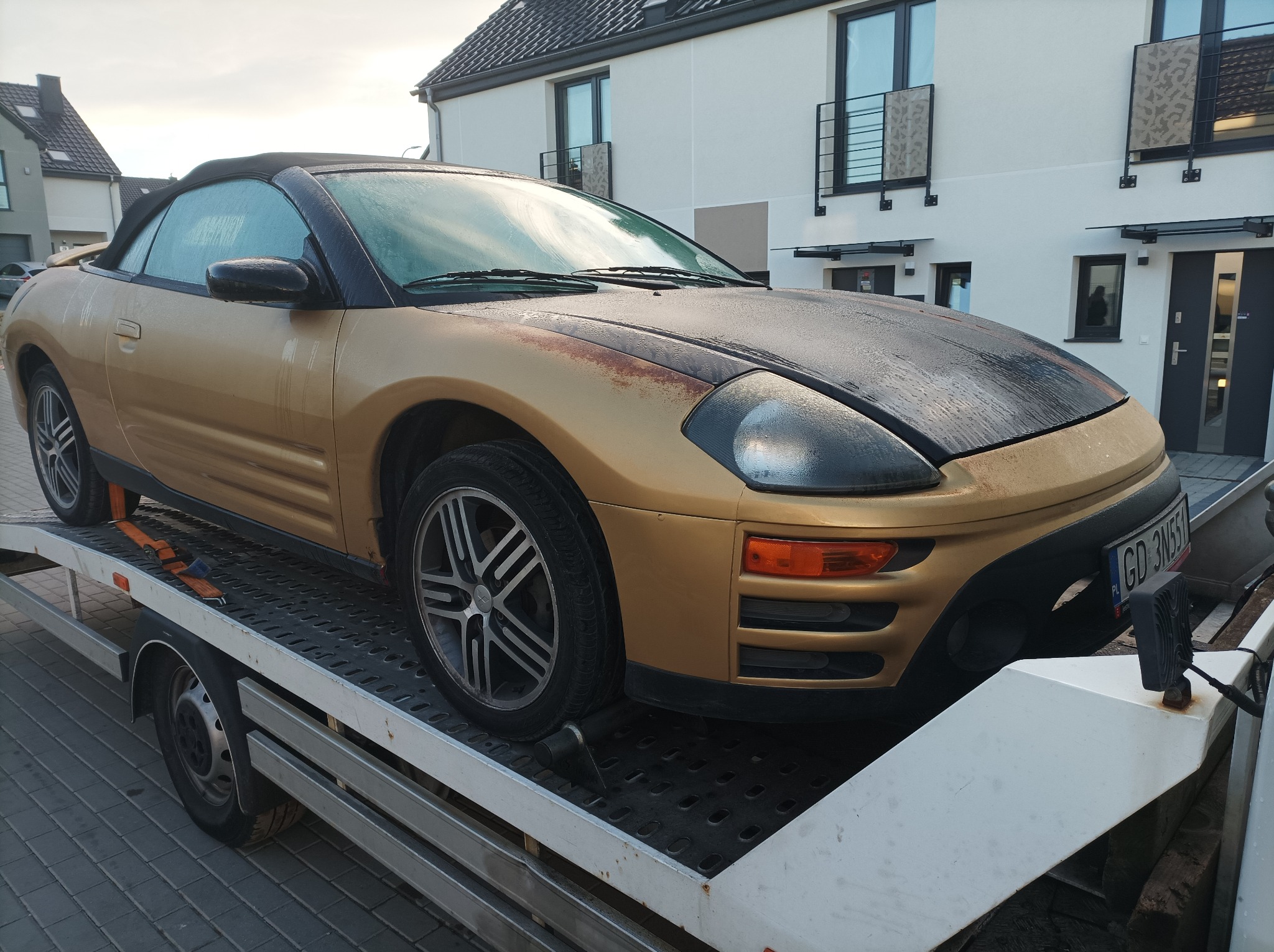 Złoty kabriolet z czarną maską, pokryty rosą, zabezpieczony pasami na platformie transportowej, na tle nowoczesnego osiedla.