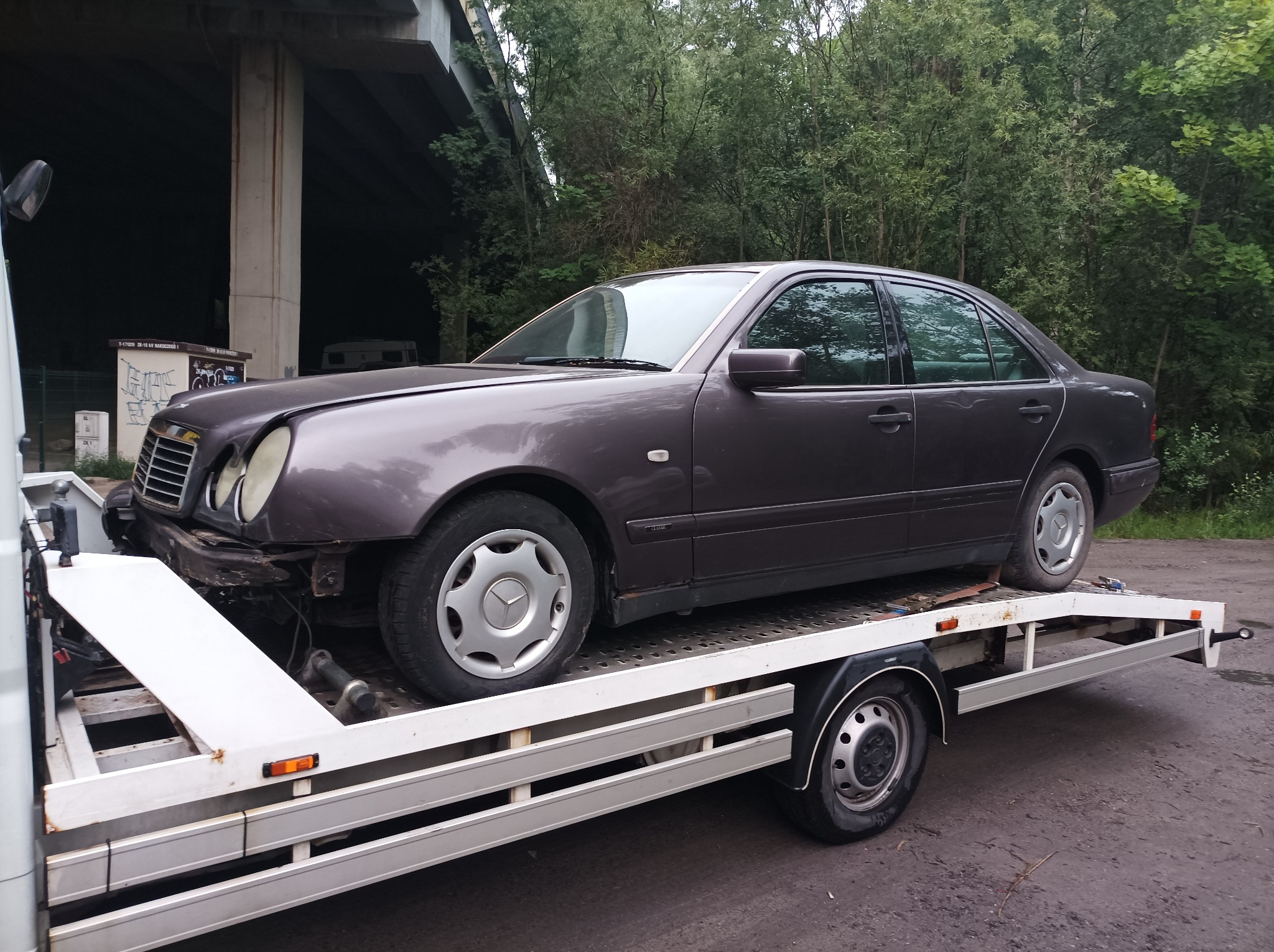 Uszkodzony, bordowy Mercedes-Benz E-klasa bez przedniego zderzaka transportowany na białej lawecie, widok z boku, w tle drzewa i filar wiaduktu.