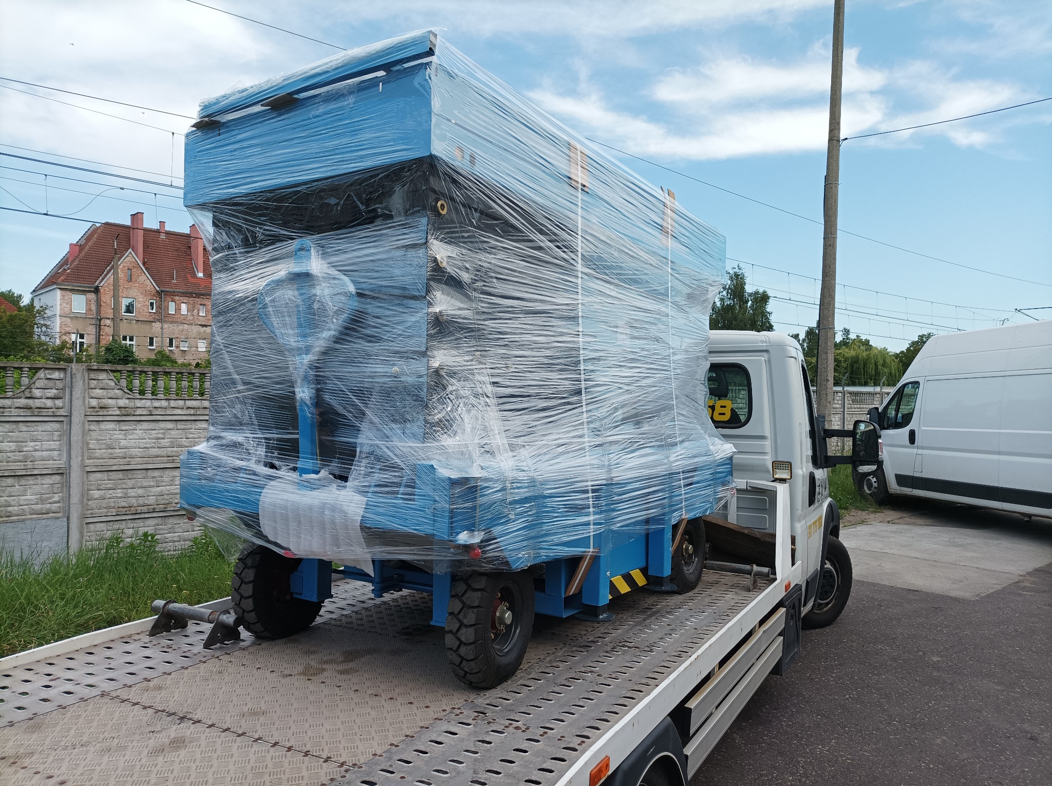Niebieska platforma robocza owinięta folią stretch, zabezpieczona na platformie transportowej, widok z tyłu, budynek mieszkalny w tle.
