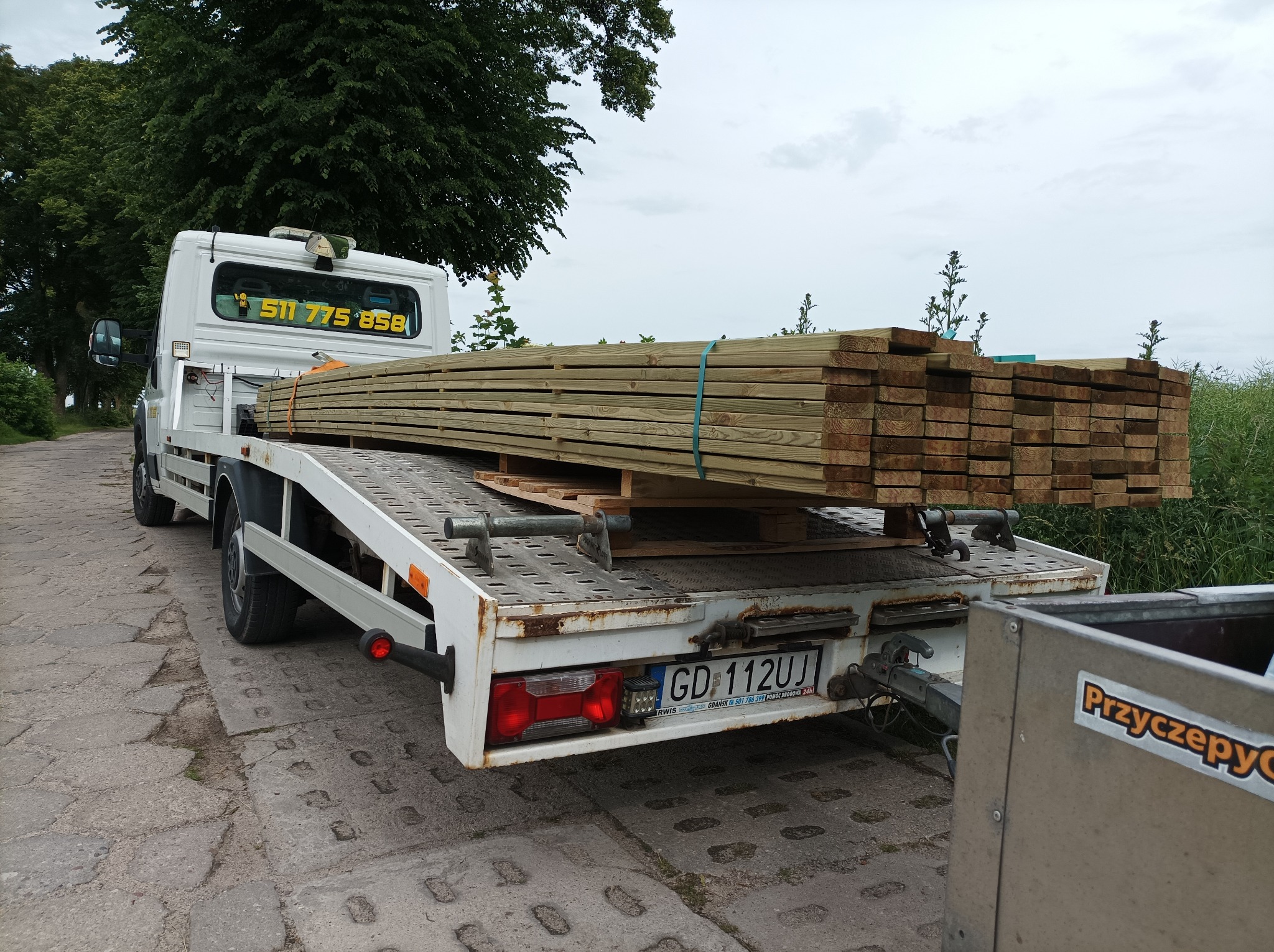 Biała platforma transportowa przewozi paletę z drewnianymi belkami, zabezpieczonymi niebieską taśmą, widok z tyłu na tle zieleni i pochmurnego nieba.