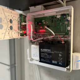 Otwarta skrzynka z elektroniką i akumulatorem alarmowym Alarmtec BP 18-12. Widoczne kable i płytki drukowane. Element systemu alarmowego wewnątrz pomieszczenia.