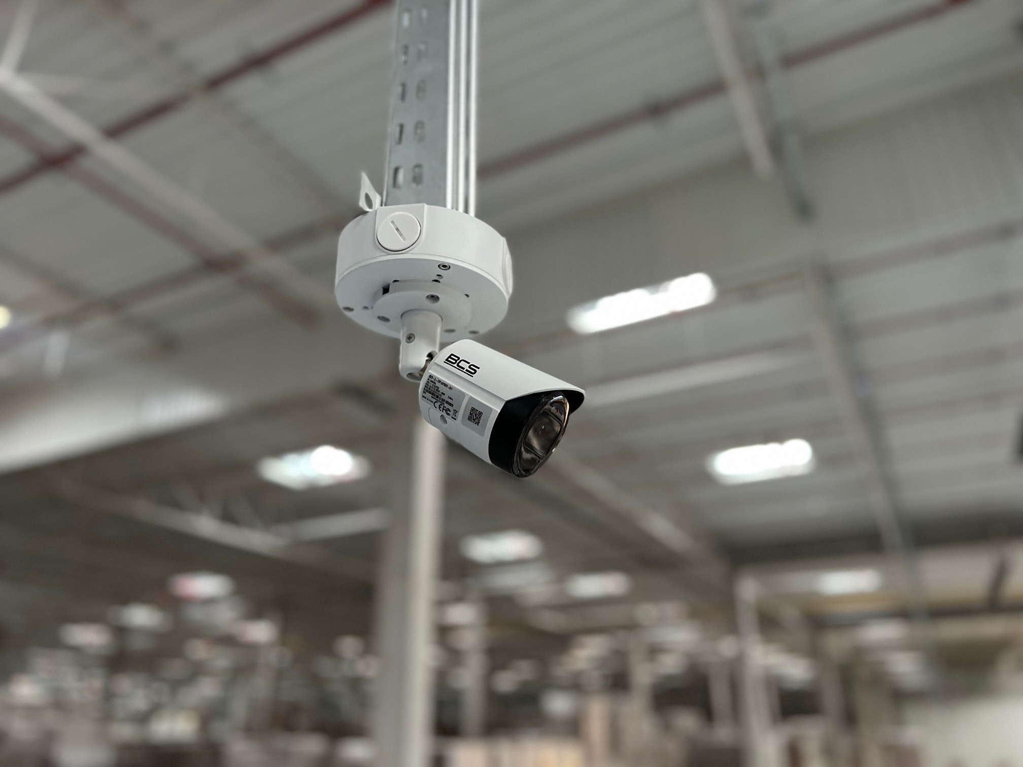 Kamera monitoringu BCS, zamontowana pod sufitem w hali magazynowej. Widoczna biała obudowa kamery i uchwyt montażowy. Tło rozmyte, ukazujące regały magazynowe.