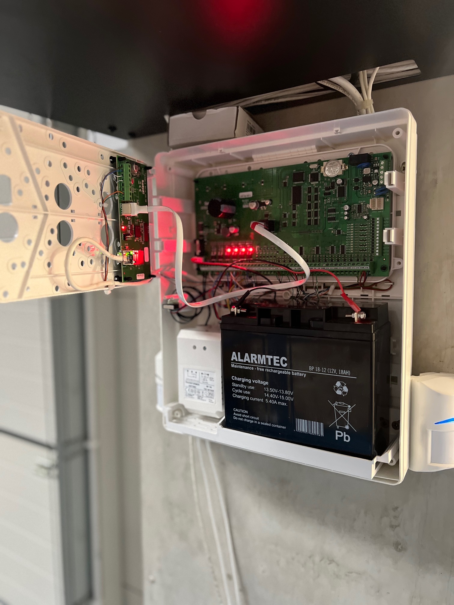 Otwarta skrzynka z elektroniką i akumulatorem alarmowym Alarmtec BP 18-12. Widoczne kable i płytki drukowane. Element systemu alarmowego wewnątrz pomieszczenia.