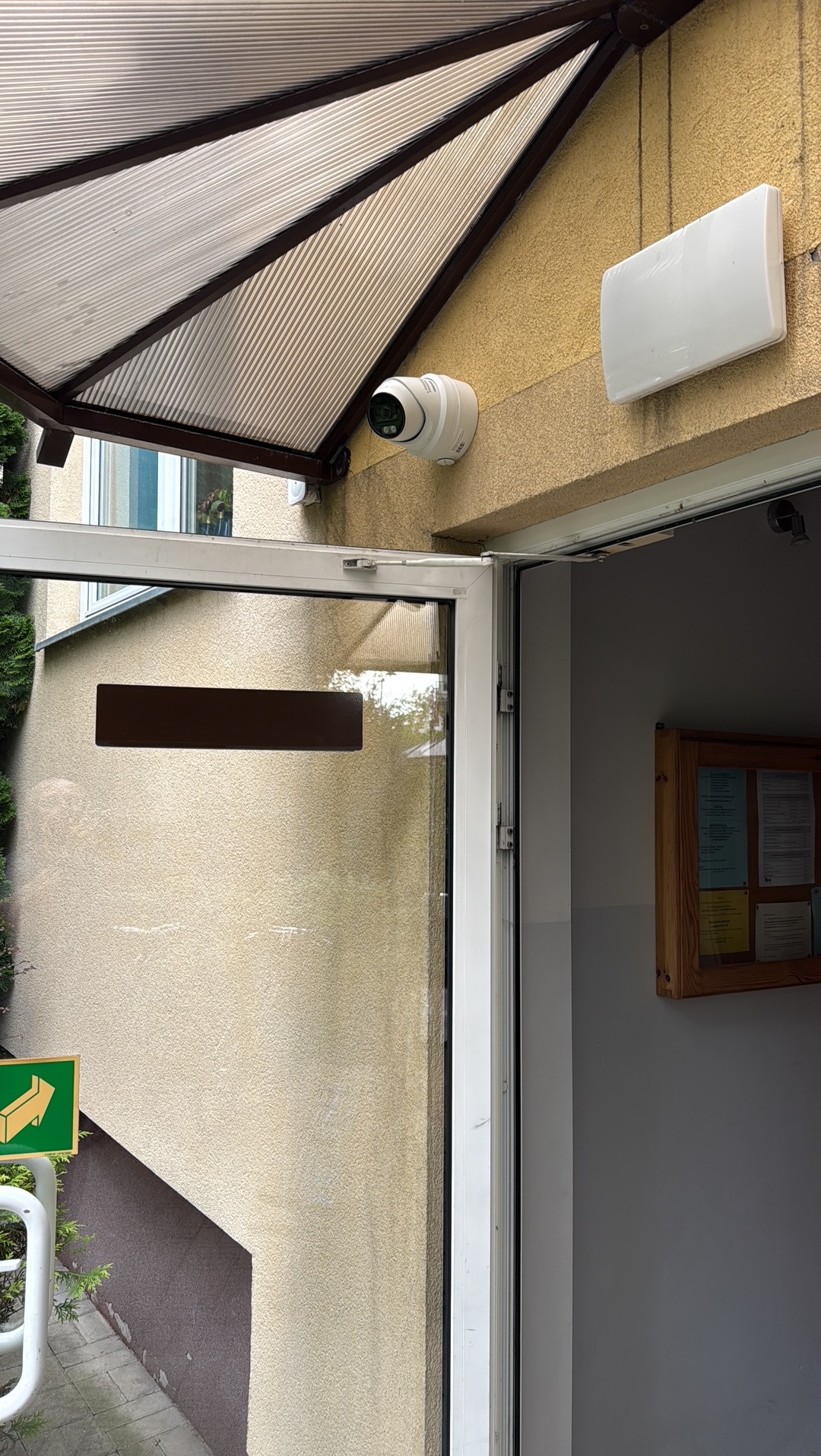 Kamera monitoringu IP zamontowana pod zadaszeniem budynku, obok biała obudowa. Widoczne wejście do budynku z otwartymi drzwiami.