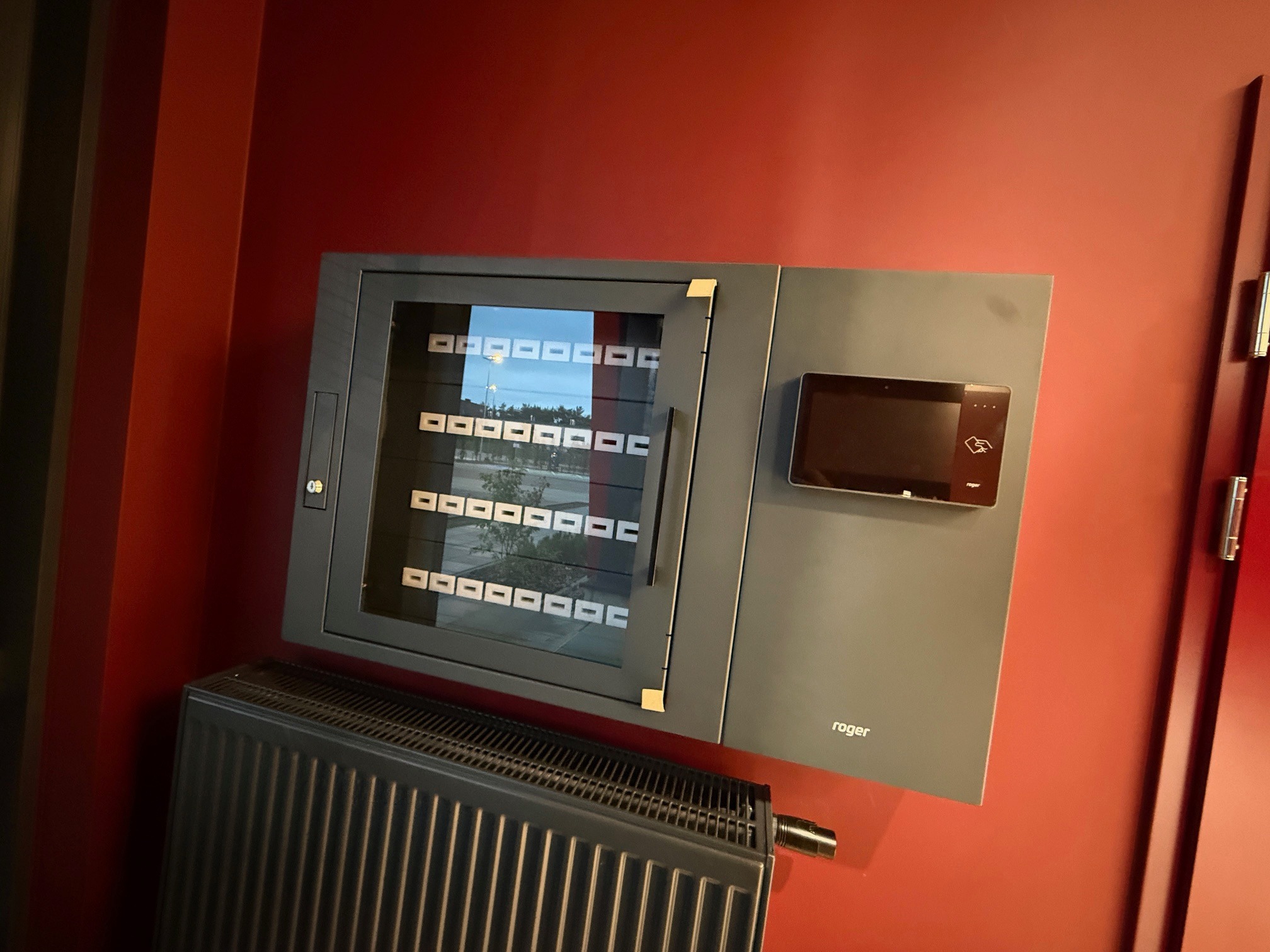 Panel kontrolny dostępu Roger z czytnikiem kart i szafką na klucze, zamontowany na czerwonej ścianie. Widoczny grzejnik pod panelem.