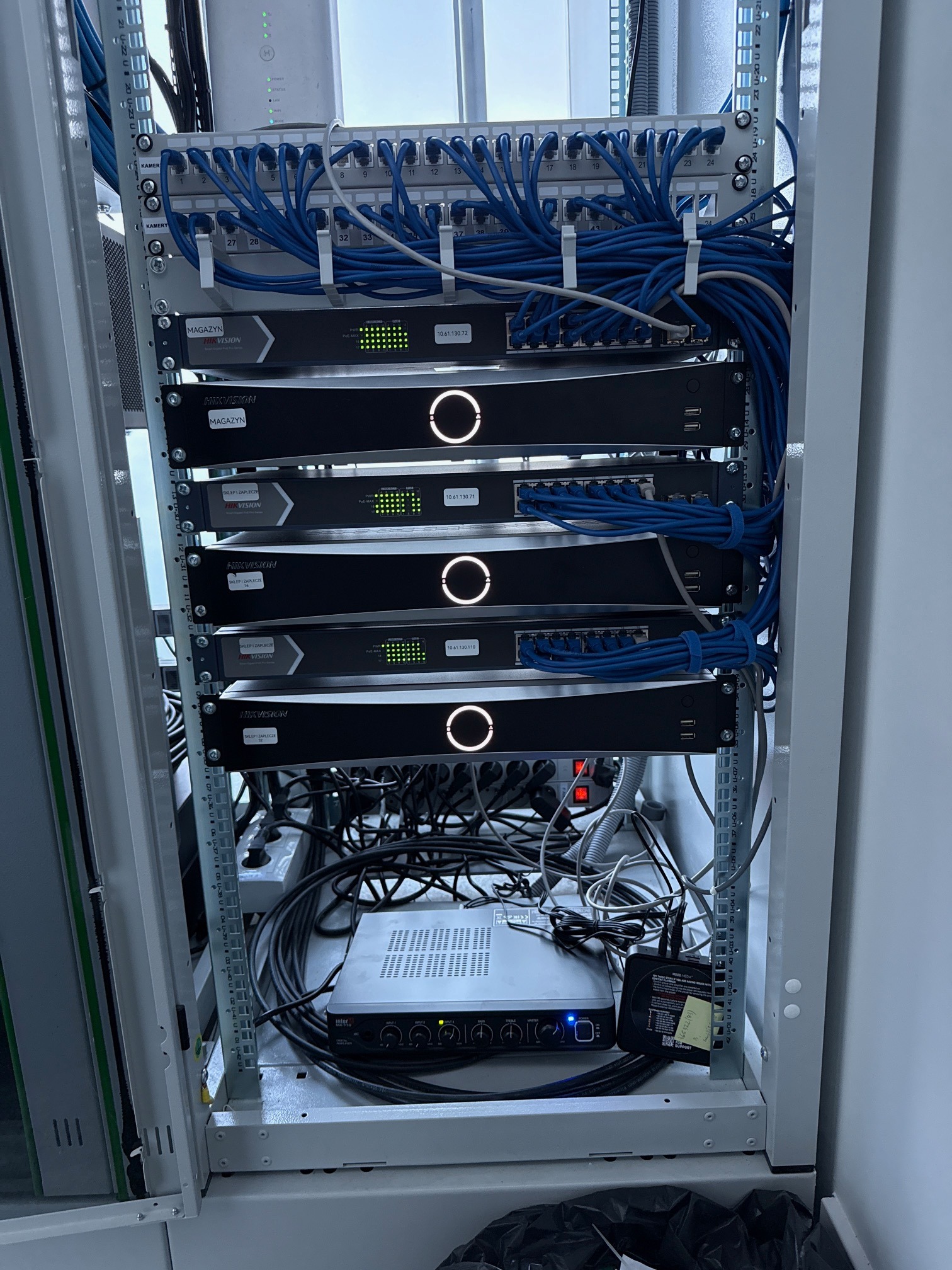 Szafa rack z zamontowanymi rejestratorami Hikvision, patch panelem i okablowaniem. Widoczne porty USB, diody sygnalizacyjne i etykiety. Estetyczne ułożenie kabli.