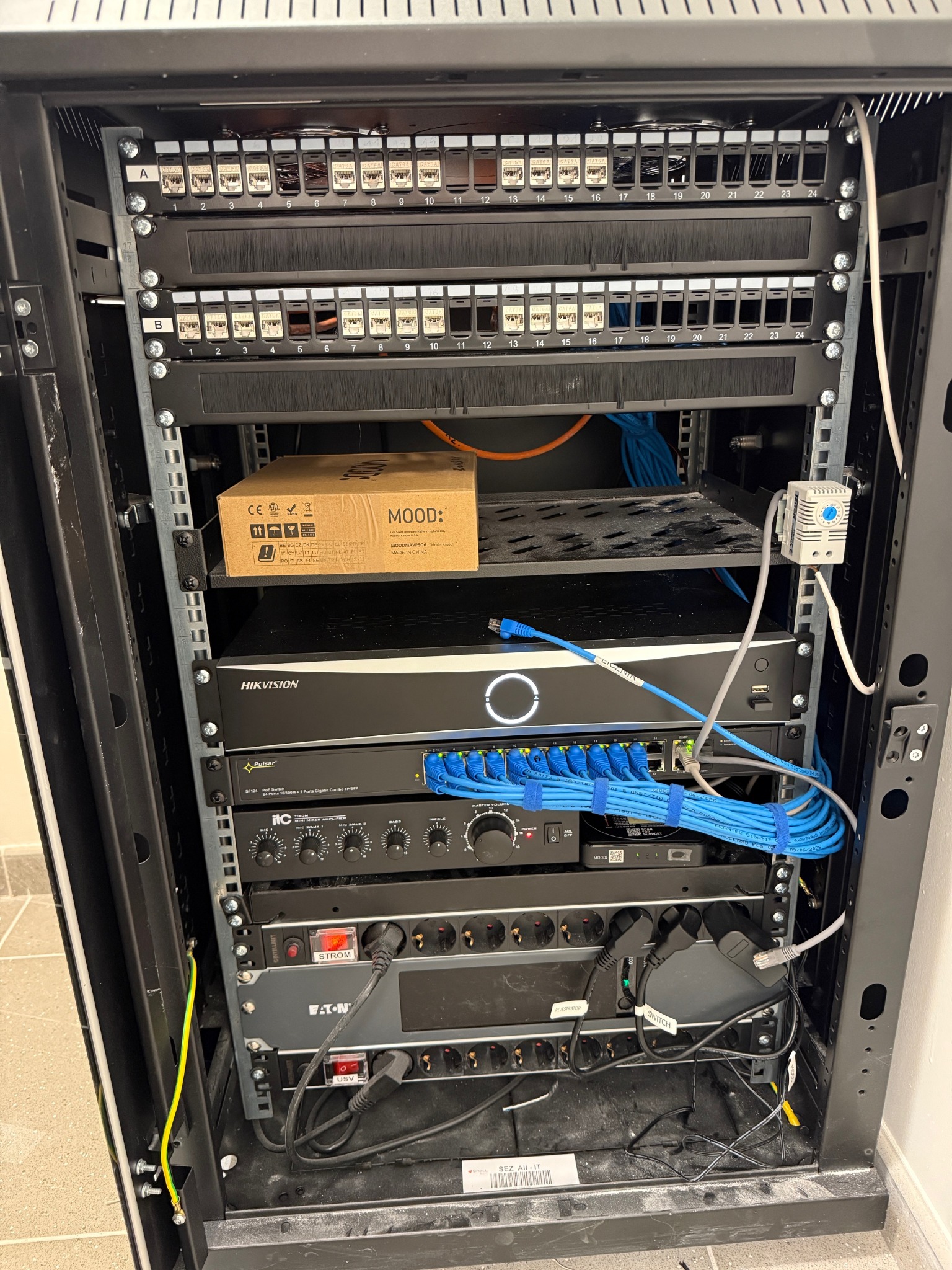 Zamknięta szafa rack z zainstalowanymi urządzeniami sieciowymi, patch panele, switch, rejestrator Hikvision, wzmacniacz ITC, zasilacz Eaton. Widoczne okablowanie.