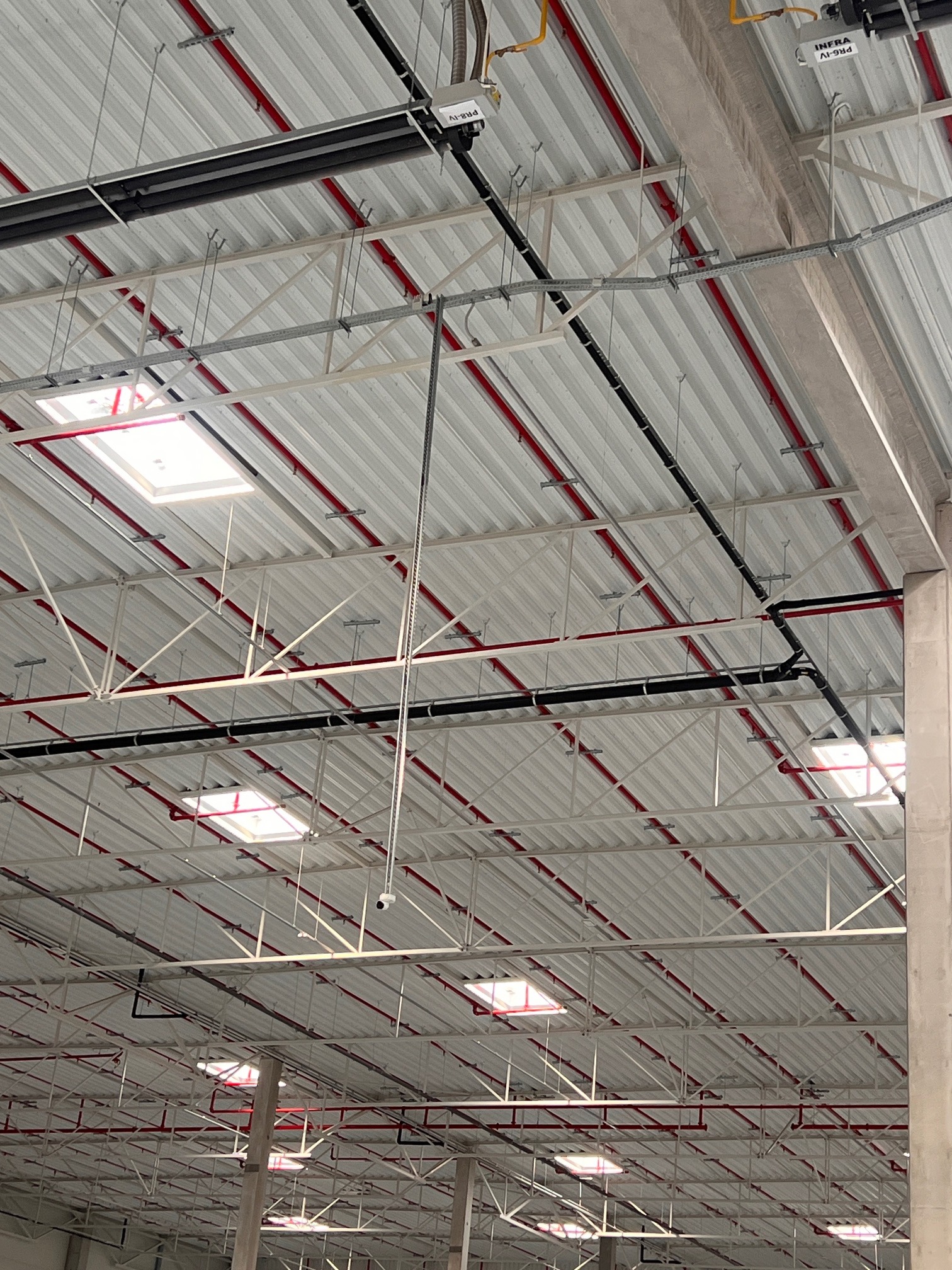 Sufit hali magazynowej z instalacją monitoringu (kamera na rurze), oświetleniem i instalacją przeciwpożarową (czerwone rury). Widok z dołu, w tle konstrukcja dachu.