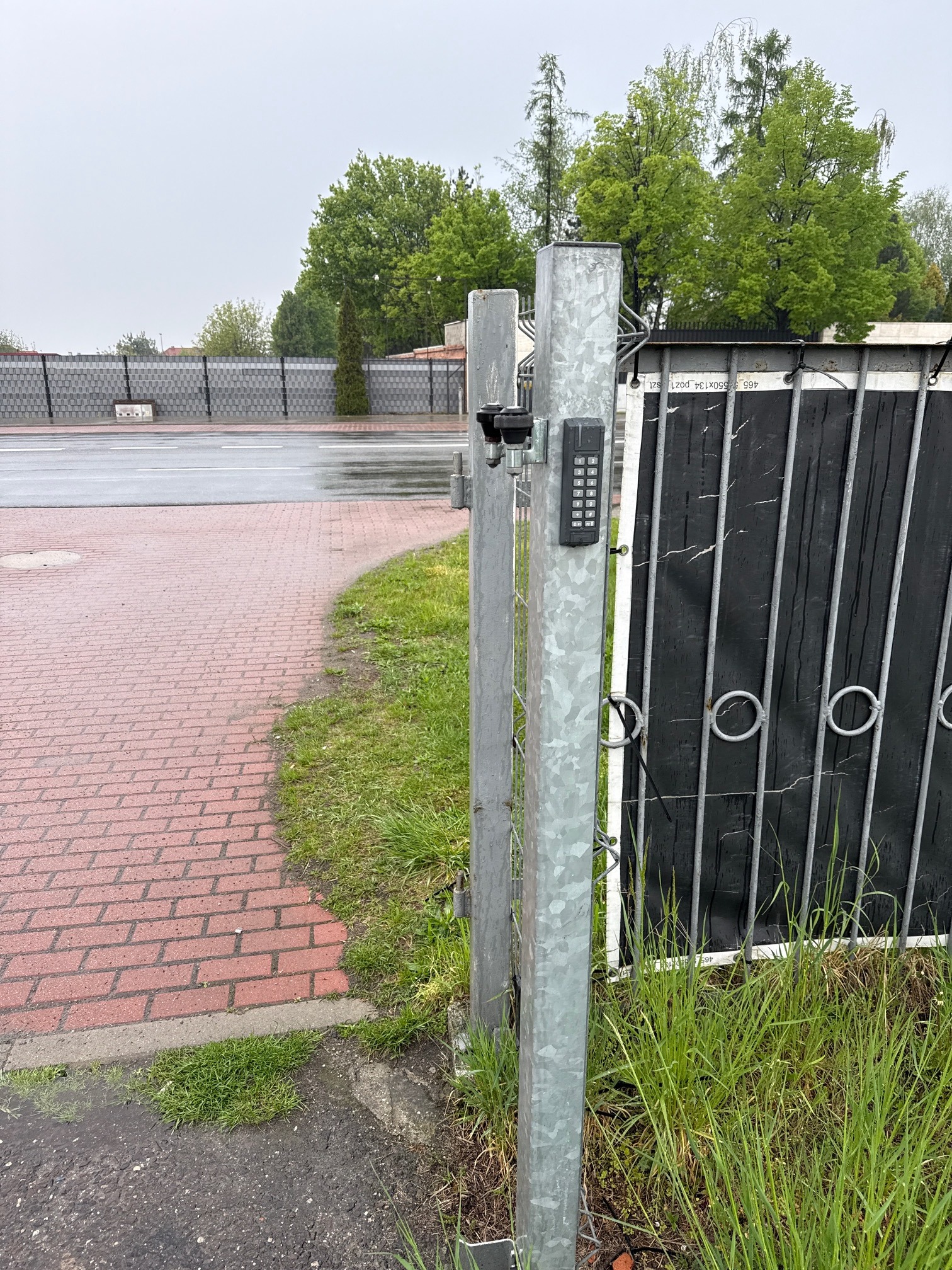 Automatyka bram wjazdowych w Warszawie: metalowa brama z panelem numerycznym i dwoma kamerami, widok z bliska na tle zieleni i kostki brukowej.