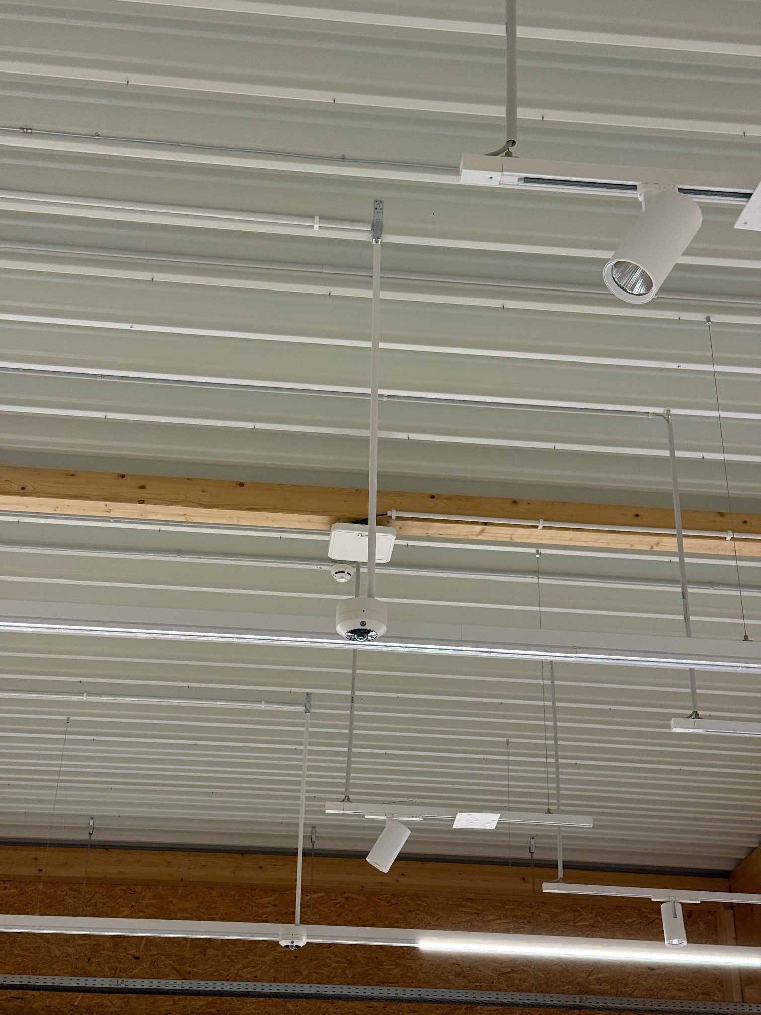 Biały sufit z blachy trapezowej z zamontowanymi lampami szynowymi, kamerą monitoringu i czujnikiem dymu. Widoczne elementy instalacji elektrycznej.