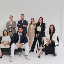 Blue Real Estate Bogacka & Gołębiowska s.c - Grupa siedmiu elegancko ubranych osób, w tym cztery kobiety i trzech mężczyzn, pozujących na białym tle w studio, z dwoma kobietami siedzącymi na czarnej kanapie i krześle.