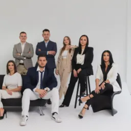 Grupa siedmiu elegancko ubranych osób, w tym cztery kobiety i trzech mężczyzn, pozujących na białym tle w studio, z dwoma kobietami siedzącymi na czarnej kanapie i krześle.