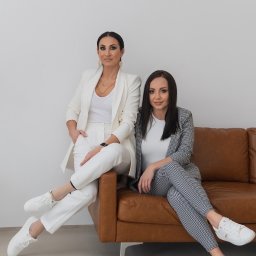Blue Real Estate Bogacka & Gołębiowska s.c - Dwie kobiety w eleganckich garniturach, siedzące na brązowej skórzanej sofie w minimalistycznym wnętrzu, prezentują profesjonalny wizerunek agencji nieruchomości.