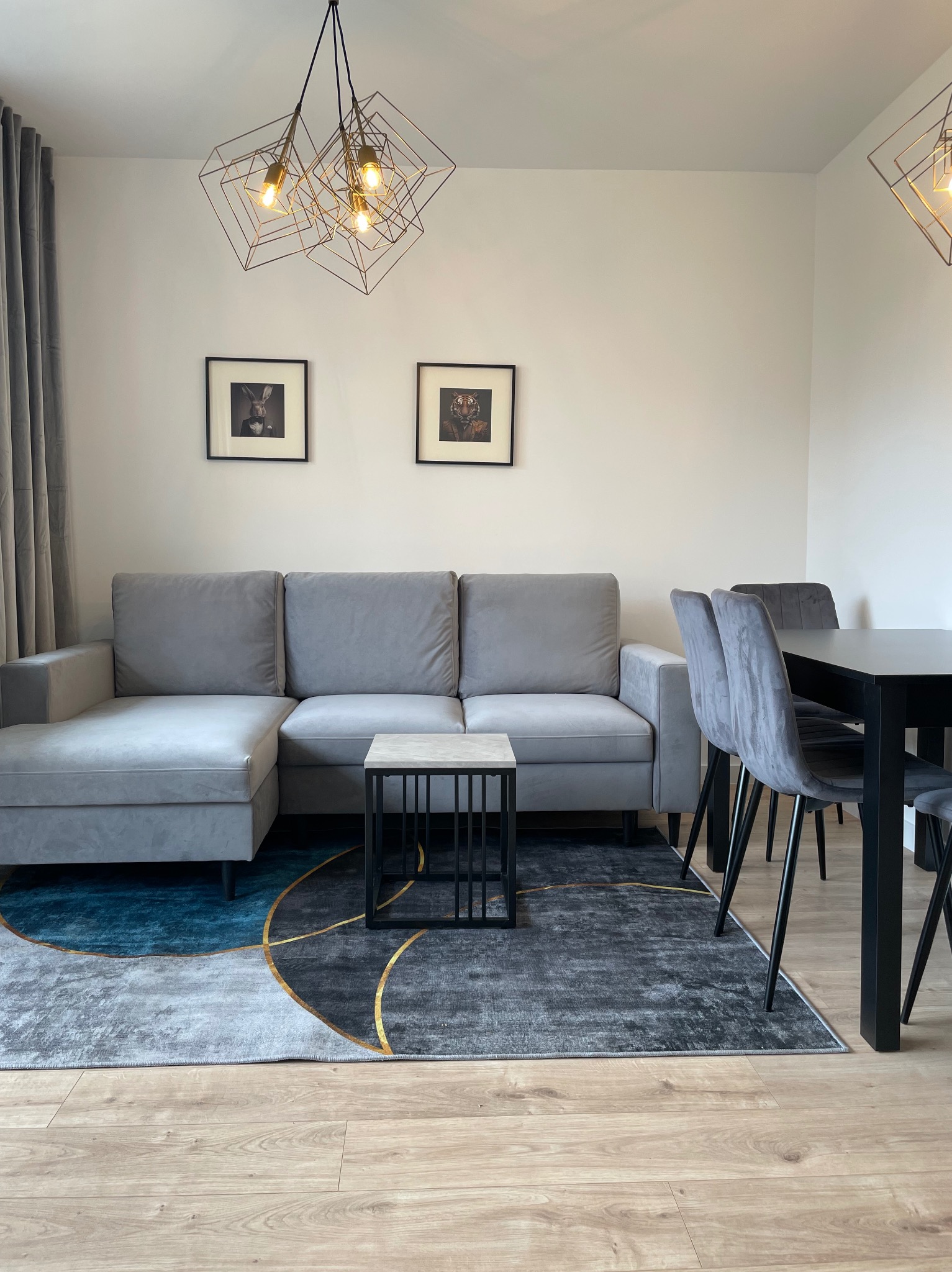 Jasne wnętrze salonu z szarą sofą narożną, stolikiem kawowym z blatem imitującym marmur, geometryczną lampą wiszącą, dywanem z abstrakcyjnym wzorem i czarnym stołem z krzesłami.