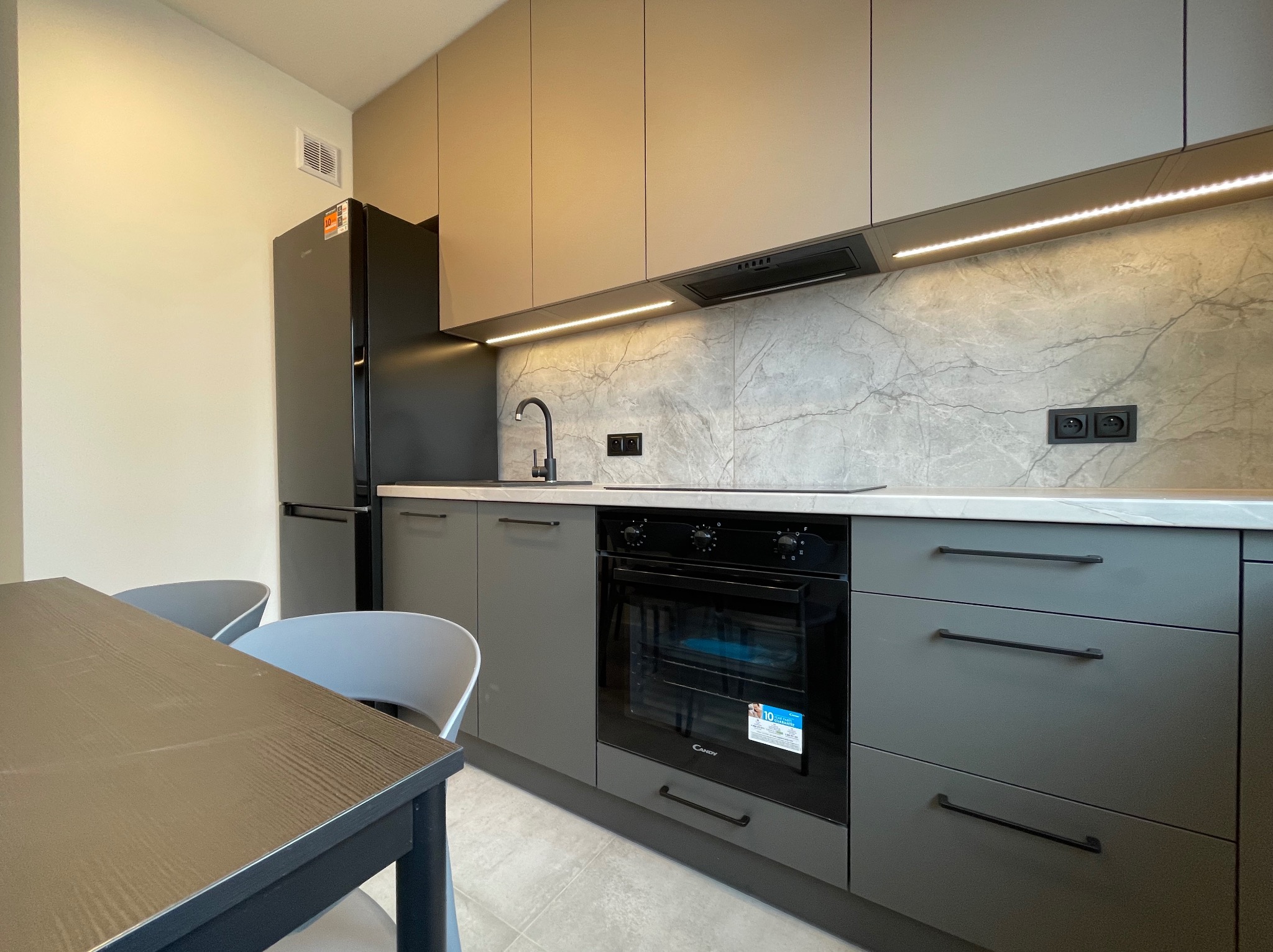 Nowoczesna kuchnia w odcieniach szarości i beżu z marmurowym backsplashiem, oświetleniem LED podszafkowym i czarnym sprzętem AGD, widoczny fragment stołu z krzesłami.