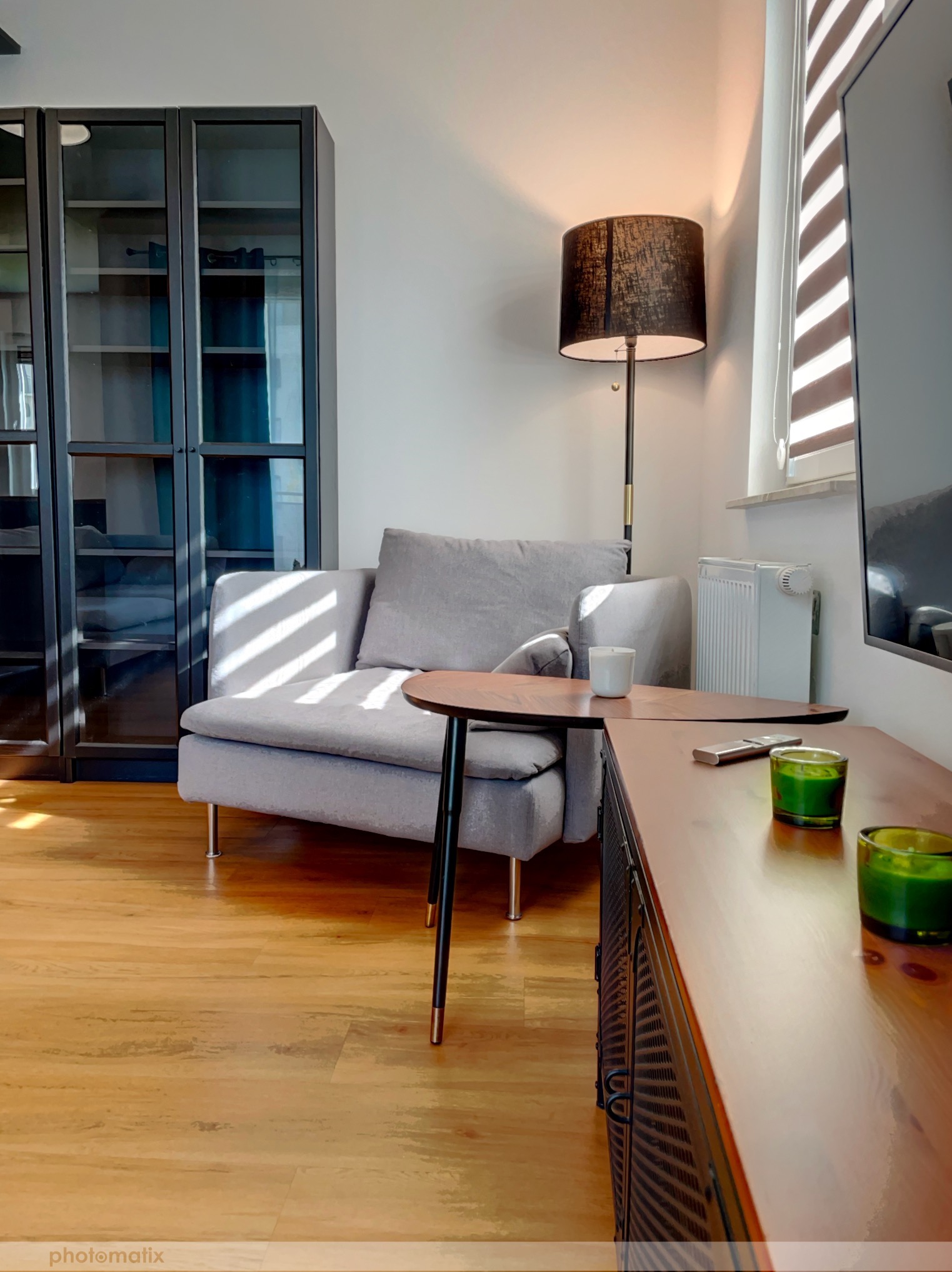 Jasnoszara sofa z poduszkami, stolik kawowy z białą świecą, drewniana szafka RTV z zielonymi świecami, czarna witryna z przeszklonymi drzwiami i lampa podłogowa z czarnym abażurem w salonie...