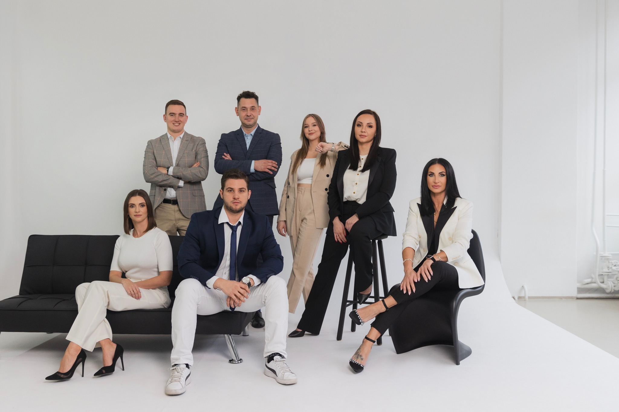 Grupa siedmiu elegancko ubranych osób, w tym cztery kobiety i trzech mężczyzn, pozujących na białym tle w studio, z dwoma kobietami siedzącymi na czarnej kanapie i krześle.