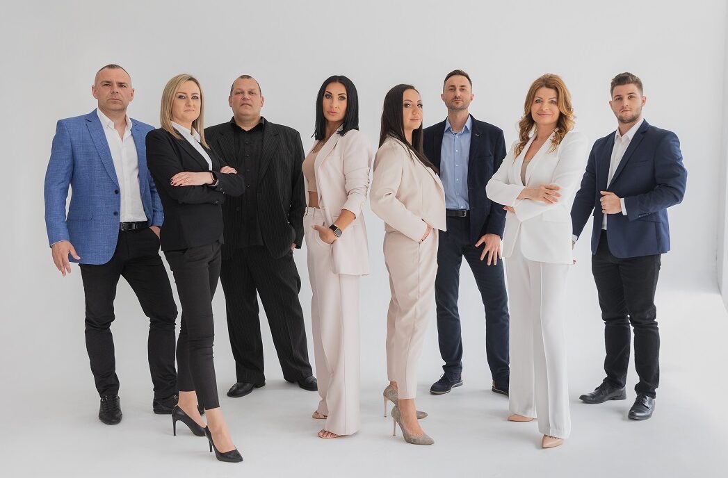 Grupa ośmiu elegancko ubranych osób, mieszany zespół w garniturach i garsonkach, stojących na białym tle, prezentujących profesjonalizm i pewność siebie, z delikatnym uśmiechem na twarzach.