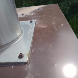 Sebbud - Metalowy komin na brązowym dachu, widoczne ślady po spawaniu i uszczelnianiu, w tle zieleń drzew.