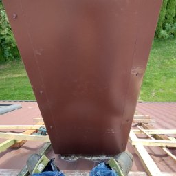 Sebbud - Widok z góry na brązowy komin blaszany w trakcie montażu na dachu pokrytym czerwoną dachówką, widoczne nogi w dżinsach i roboczych butach, fragment zielonego trawnika w tle.