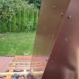 Widok z góry na fragment dachu z brązową obróbką blacharską, częściowo pokryty czerwoną dachówką i drewnianą konstrukcją, z narzędziem akumulatorowym leżącym na belce, a w tle zielony trawnik...