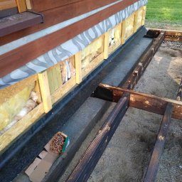 Sebbud - Budowa domu szkieletowego: widok na fundament z izolacją, fragment ściany z ociepleniem i folią, oraz karton z gwoździami na piasku.