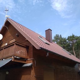 Sebbud - Brązowy dom szkieletowy z drewnianą elewacją i dachem z brązowej dachówki, widoczne okna dachowe, komin, antena satelitarna i balkon z drewnianą balustradą na tle błękitnego nieba z chmurami.