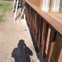 Sebbud - Nowy taras z drewnianą balustradą przy brązowym domu, widoczna drabina i materiały budowlane, cień fotografa na pierwszym planie.