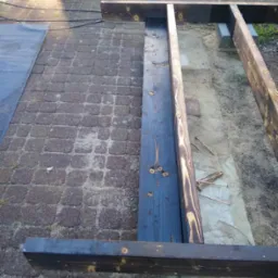 Konstrukcja ramy tarasu drewnianego w trakcie budowy, widoczne impregnowane deski, betonowe bloczki, ceglana kostka brukowa i rozsypane wkręty.