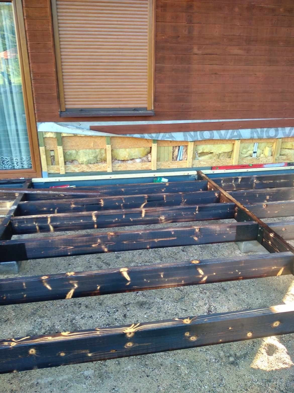 Konstrukcja nośna z ciemnego drewna, prawdopodobnie pod taras, widoczna izolacja ściany budynku w tle.