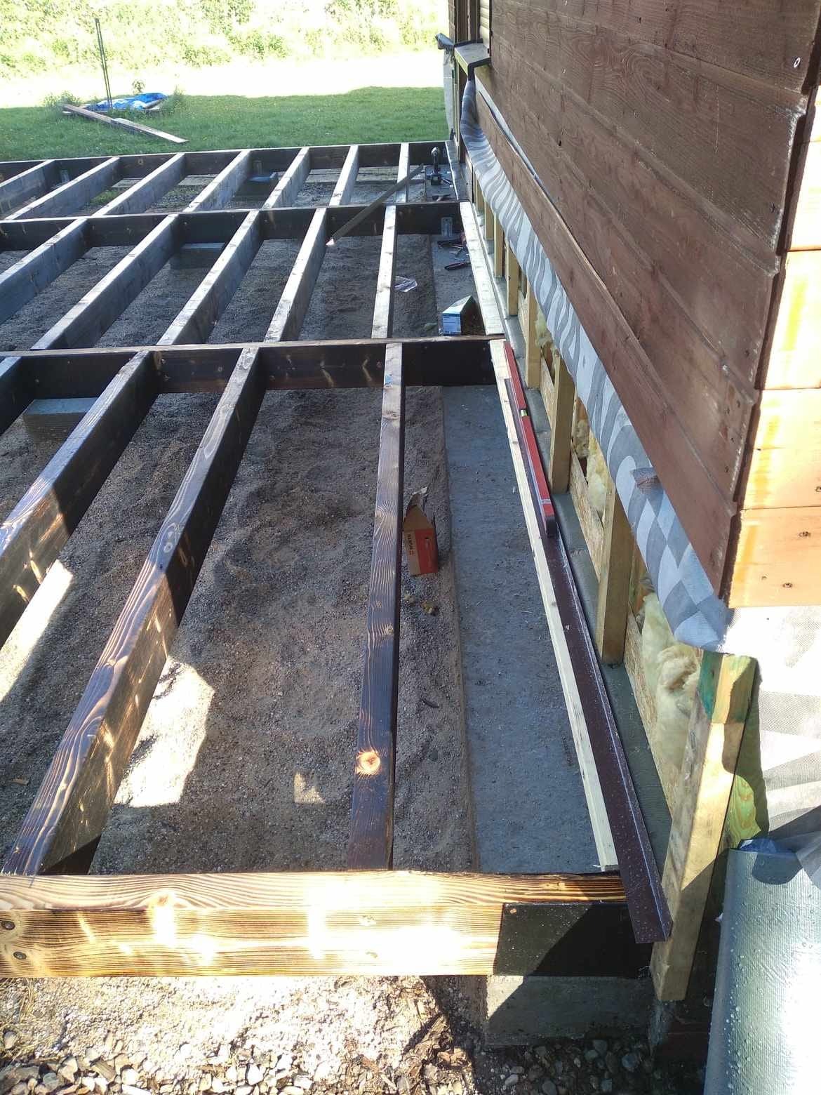 Konstrukcja drewniana pod przyszły taras przylegająca do ściany budynku, widoczne legary, podłoże z piasku i częściowo z betonu, poziomica na ścianie.