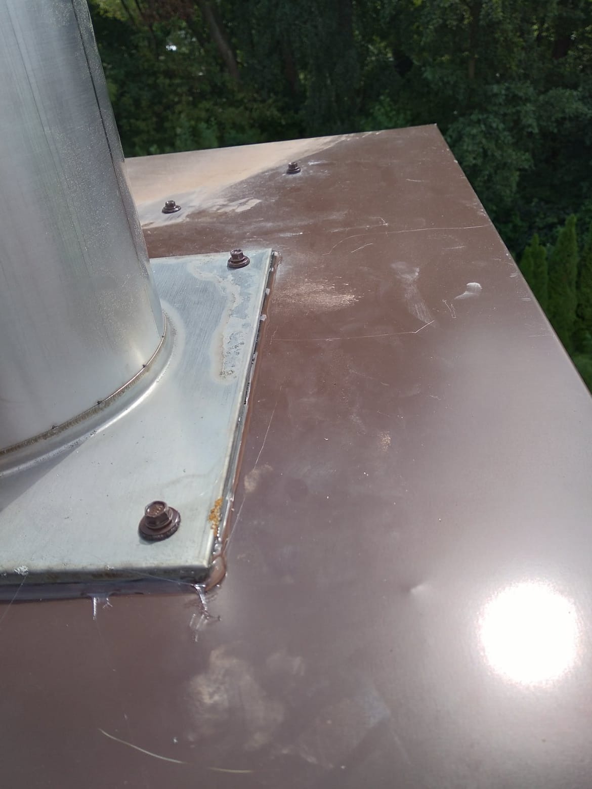 Metalowy komin na brązowym dachu, widoczne ślady po spawaniu i uszczelnianiu, w tle zieleń drzew.