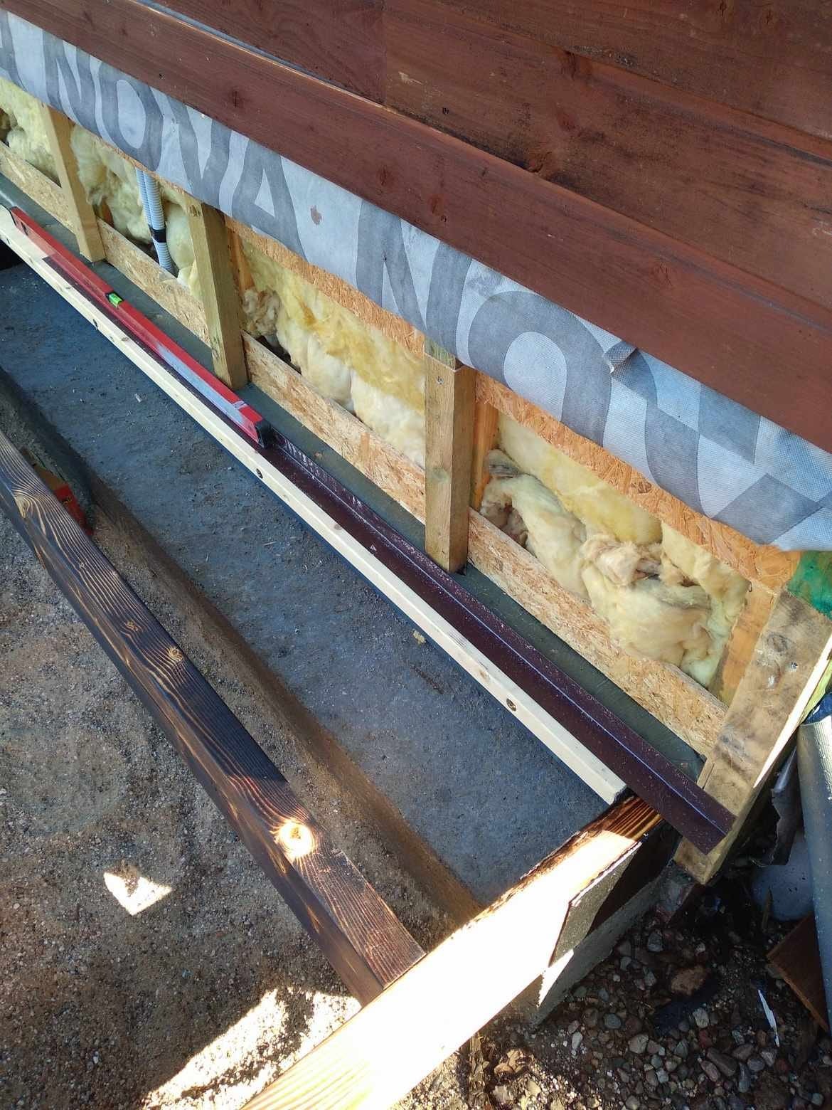 Fragment konstrukcji drewnianej z izolacją z wełny mineralnej, zabezpieczonej folią, widoczny poziomica i pomalowana listwa, etap budowy.