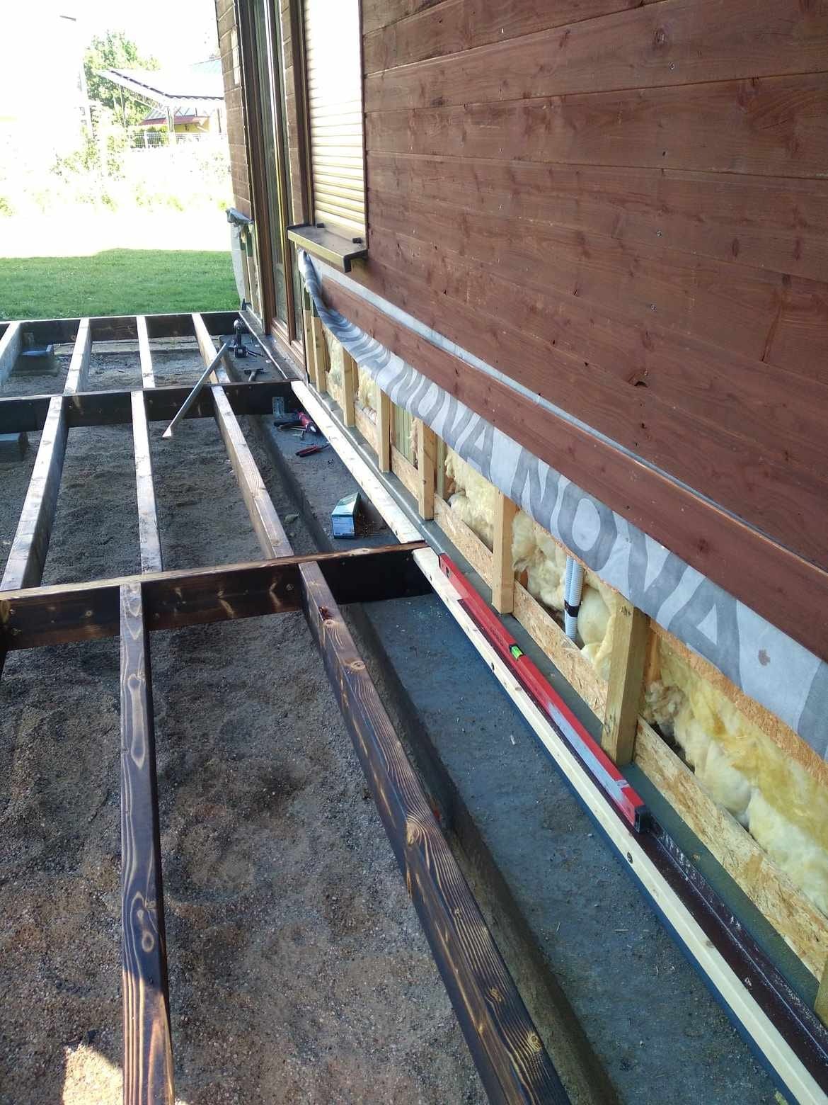 Konstrukcja nośna z belek pod przyszły taras, widoczne izolacja ściany budynku, poziomica i narzędzia.