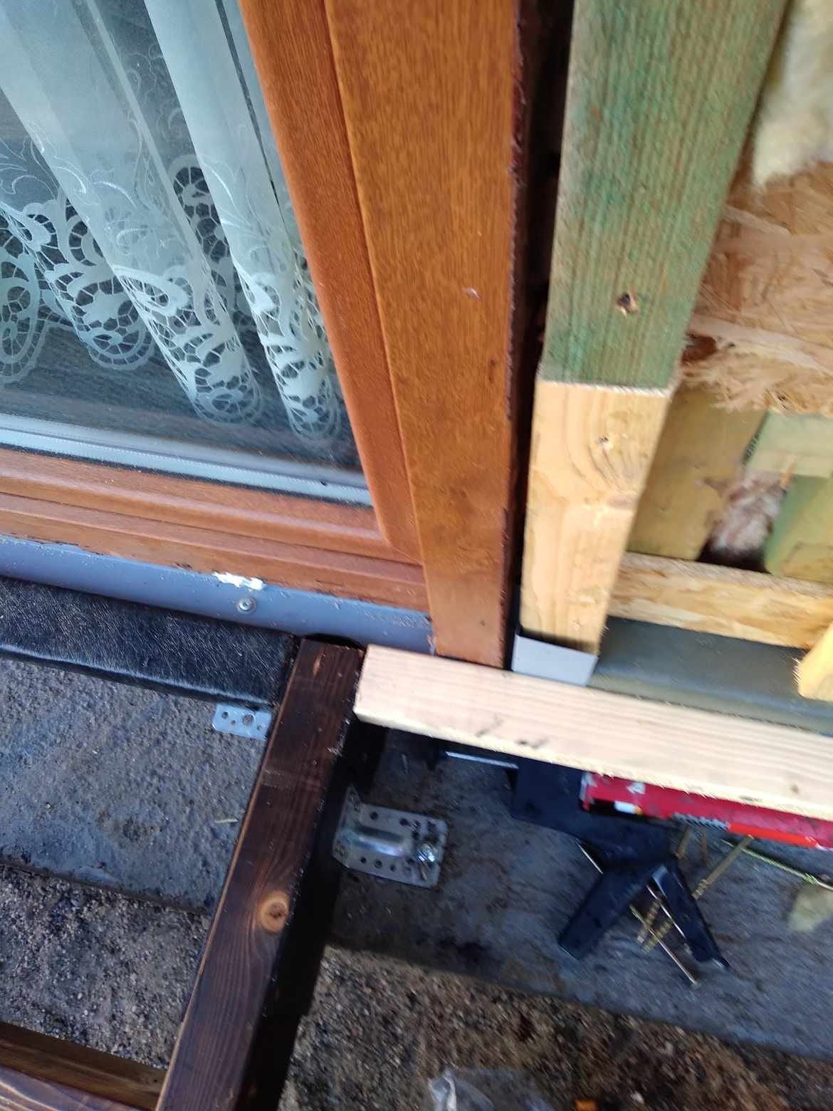 Fragment konstrukcji domu szkieletowego w trakcie budowy z widocznym oknem i izolacją, mocowania metalowe oraz narzędzia budowlane w tle.