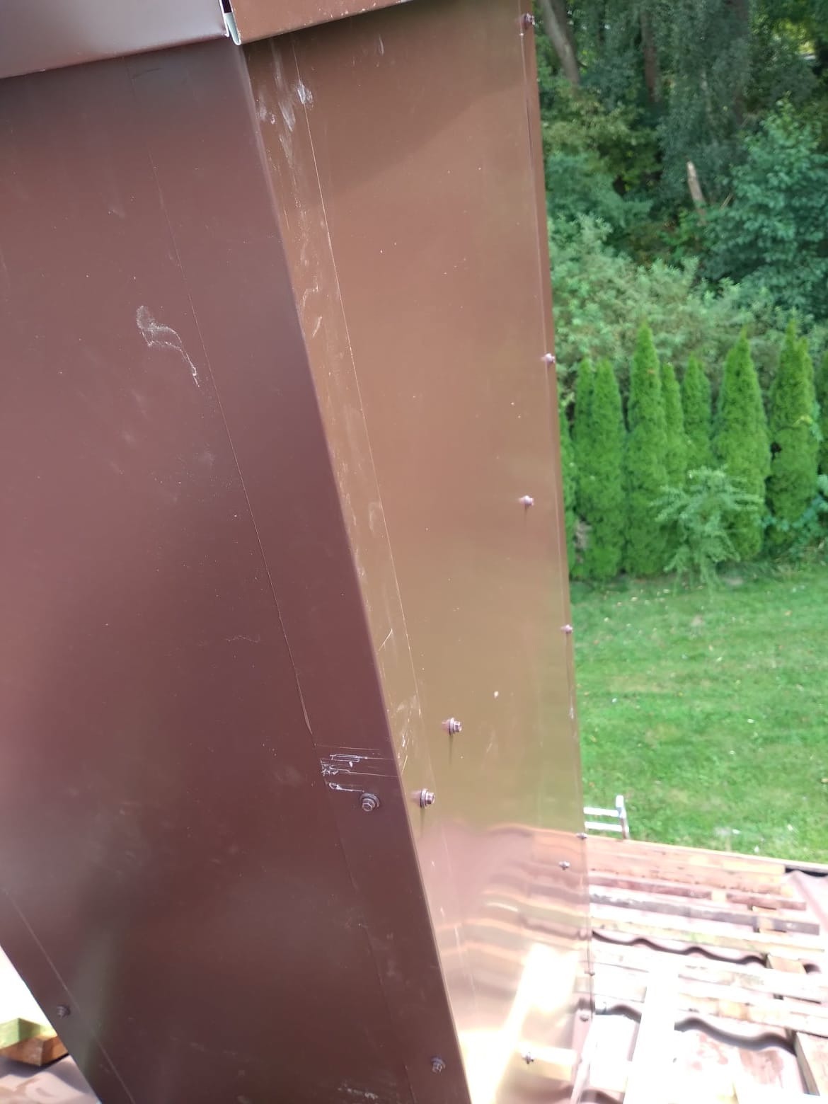 Brązowa obróbka blacharska komina, mocowana na nity, widoczna na tle zielonego trawnika i drzew.