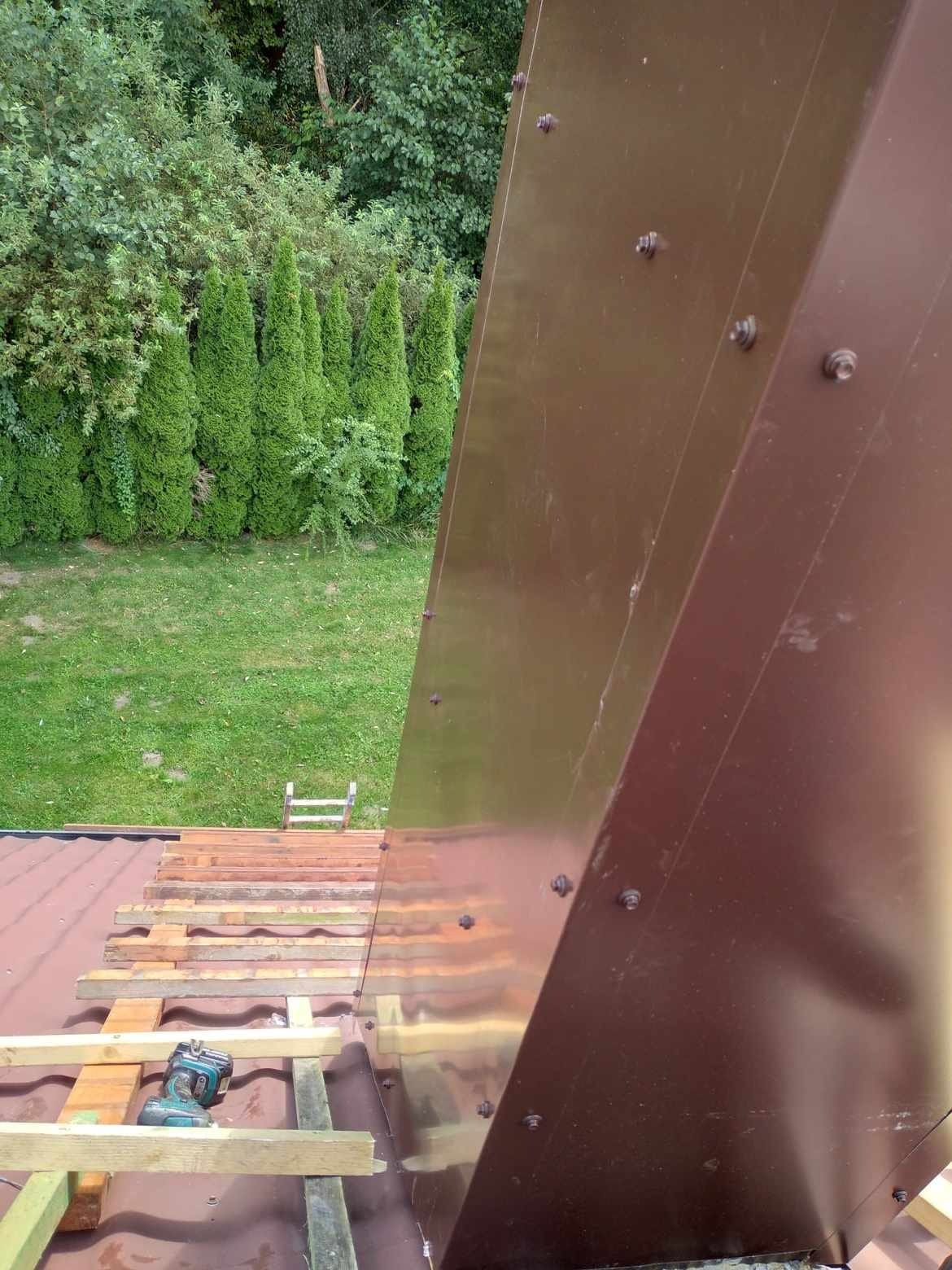 Widok z góry na fragment dachu z brązową obróbką blacharską, częściowo pokryty czerwoną dachówką i drewnianą konstrukcją, z narzędziem akumulatorowym leżącym na belce, a w tle zielony trawnik...