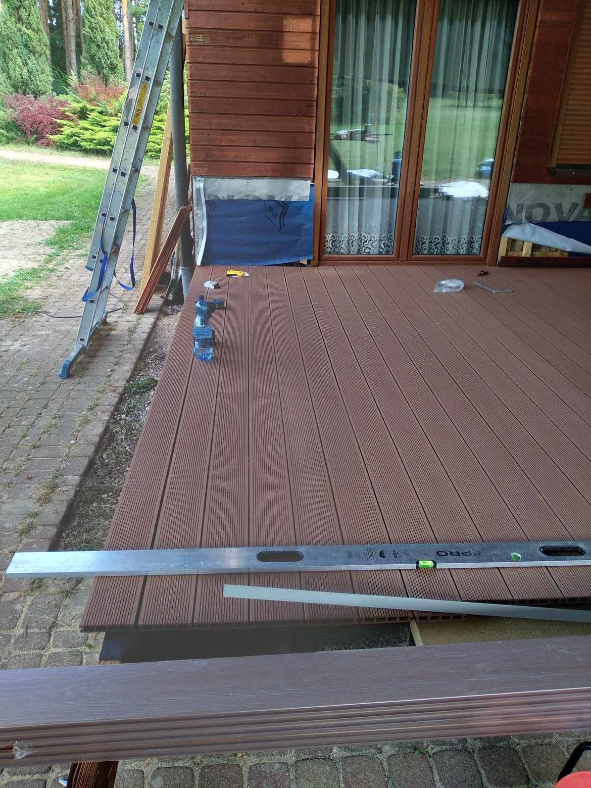 Montaż brązowego tarasu kompozytowego w Dębnie, widoczne deski, poziomica, butelki z wodą, drabina i fragment domu z oknami.
