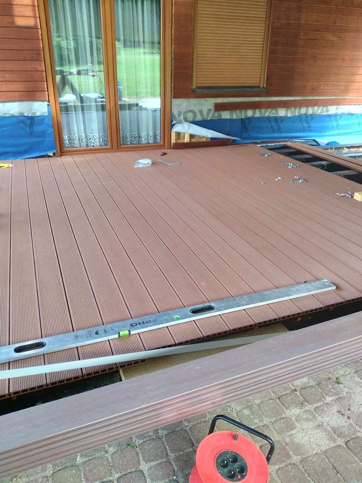 Montaż brązowego tarasu kompozytowego, widoczna poziomica i częściowo ułożone deski, pod spodem niebieska folia izolacyjna, w tle drewniana elewacja domu z oknem.