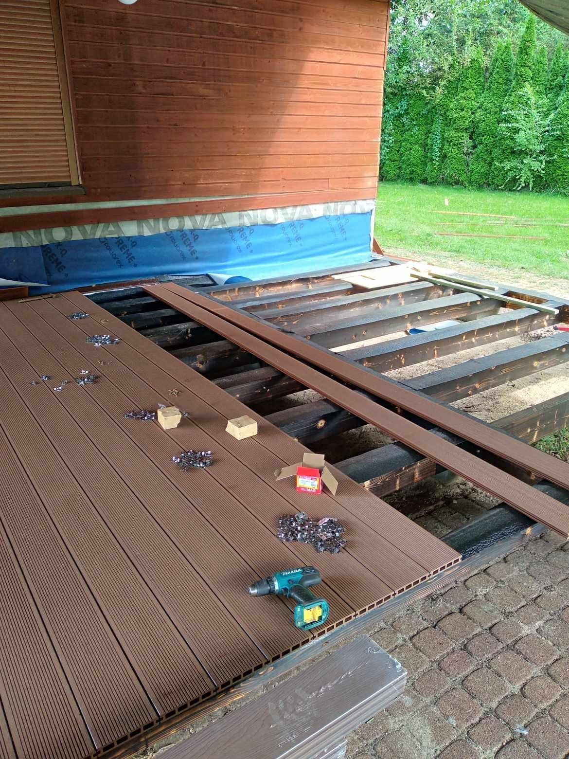 Montaż brązowego tarasu kompozytowego: deski, legary, klipsy montażowe, wkrętarka akumulatorowa Makita, widok na częściowo ułożoną konstrukcję z drewnianym domkiem w tle.