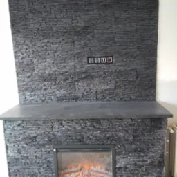 Kominek elektryczny obłożony szarą, kamienną okładziną z widocznymi gniazdkami elektrycznymi powyżej paleniska. Całość w szaro-czarnej tonacji.