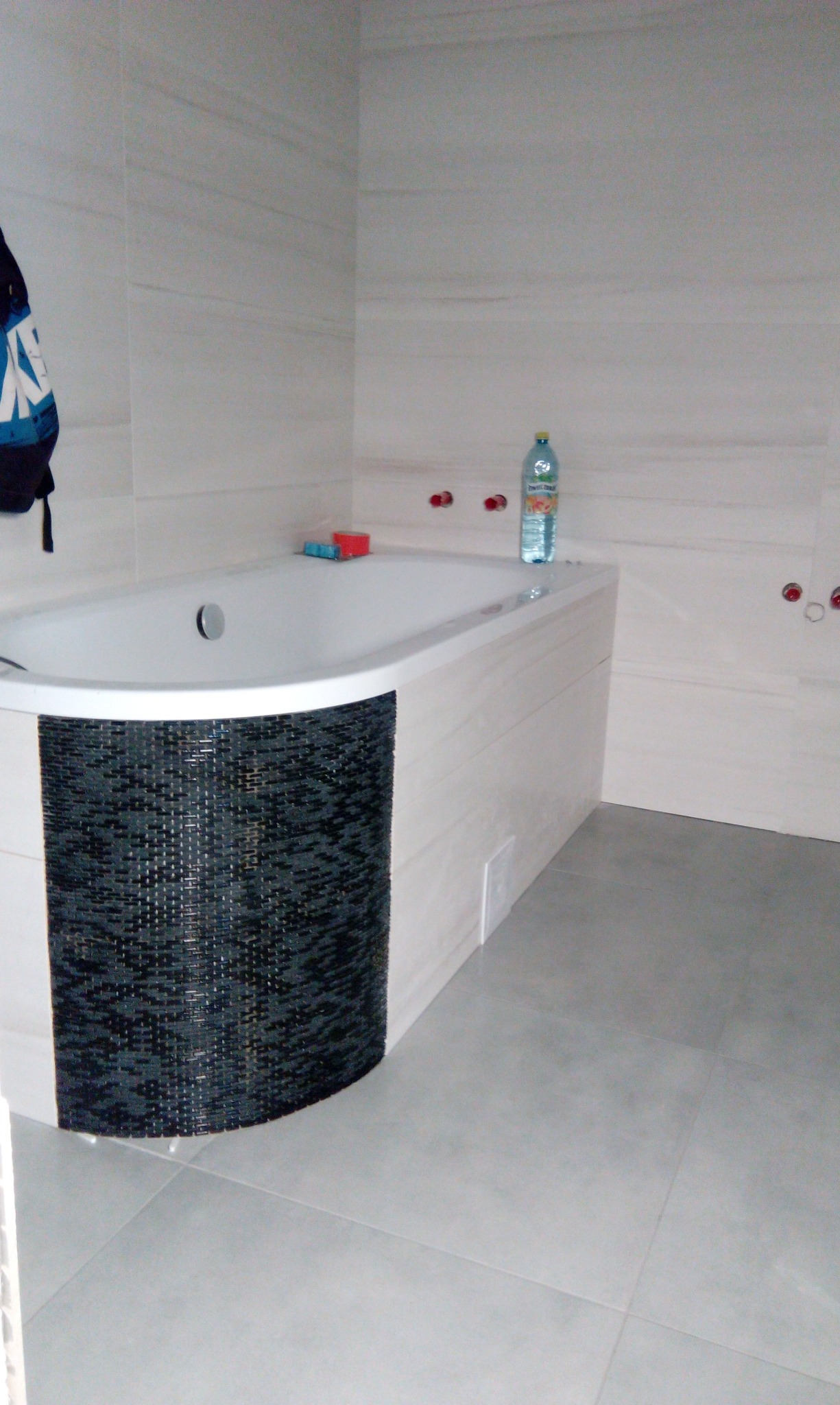 Nowoczesna łazienka z wanną w zabudowie mozaiką, płytki gresowe na ścianach i podłodze. Widoczne przyłącza hydrauliczne i butelka wody mineralnej.