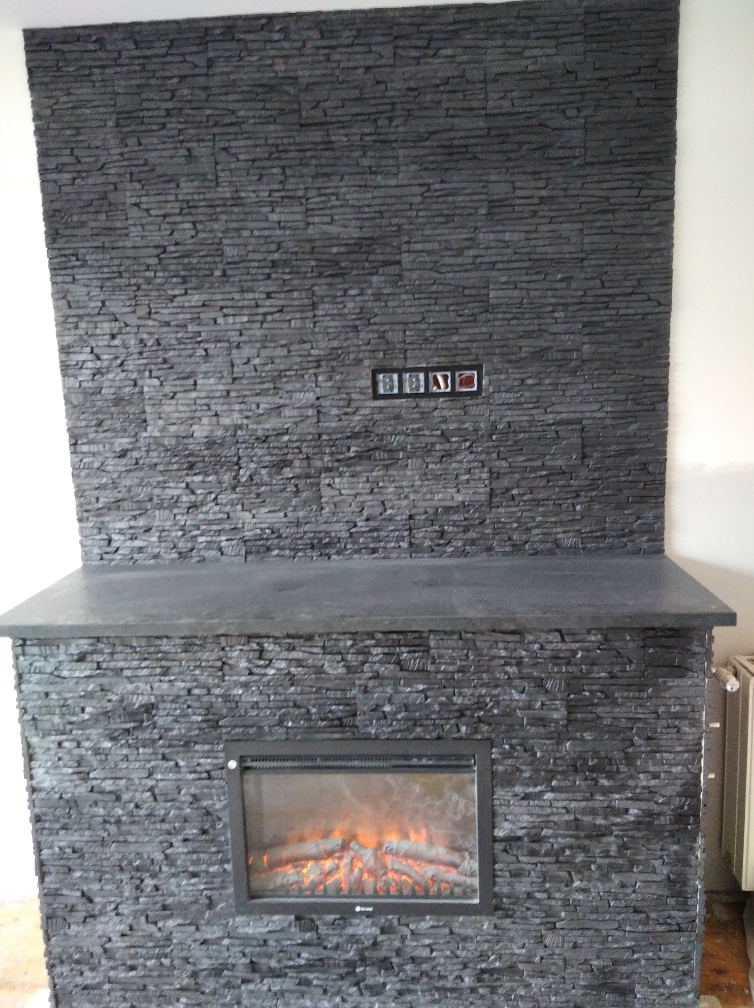 Kominek elektryczny obłożony szarą, kamienną okładziną z widocznymi gniazdkami elektrycznymi powyżej paleniska. Całość w szaro-czarnej tonacji.