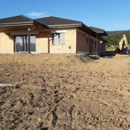 Budynek w stanie surowym otwartym z widoczną konstrukcją dachu i pustakami ceramicznymi, z żółtą koparką w tle na nierównym terenie.