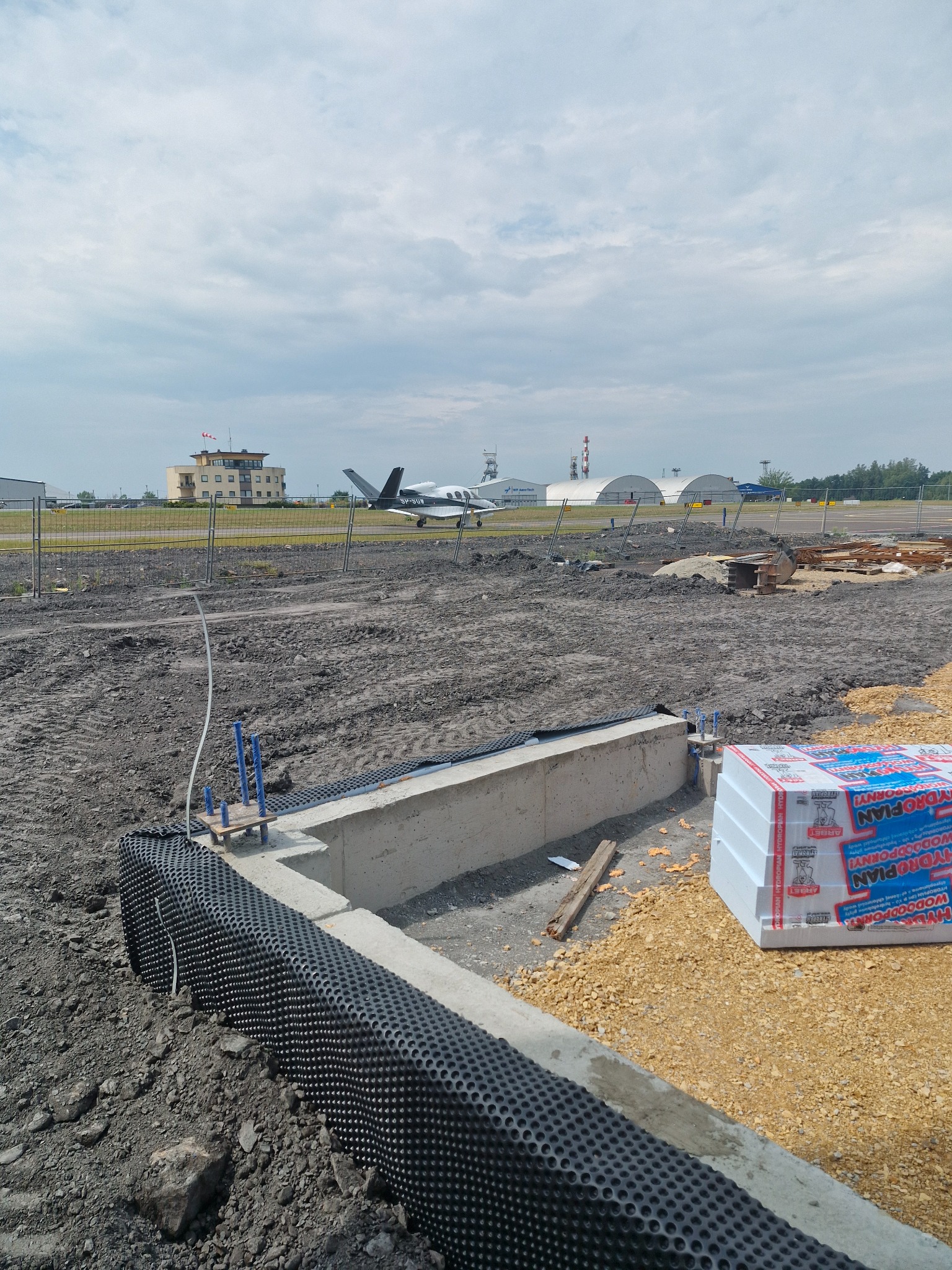 Wykop fundamentowy z hydroizolacją w postaci czarnej folii kubełkowej, widoczne wystające pręty zbrojeniowe, obok stos płyt styropianowych z niebieskim nadrukiem, w tle lotnisko z samolotem.